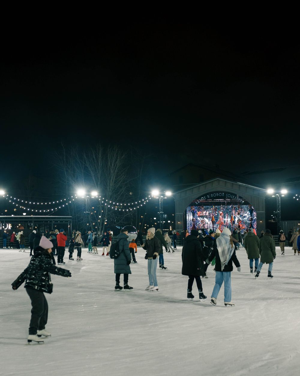 Skaters on an outdoor ice rink at night under festoon lights
