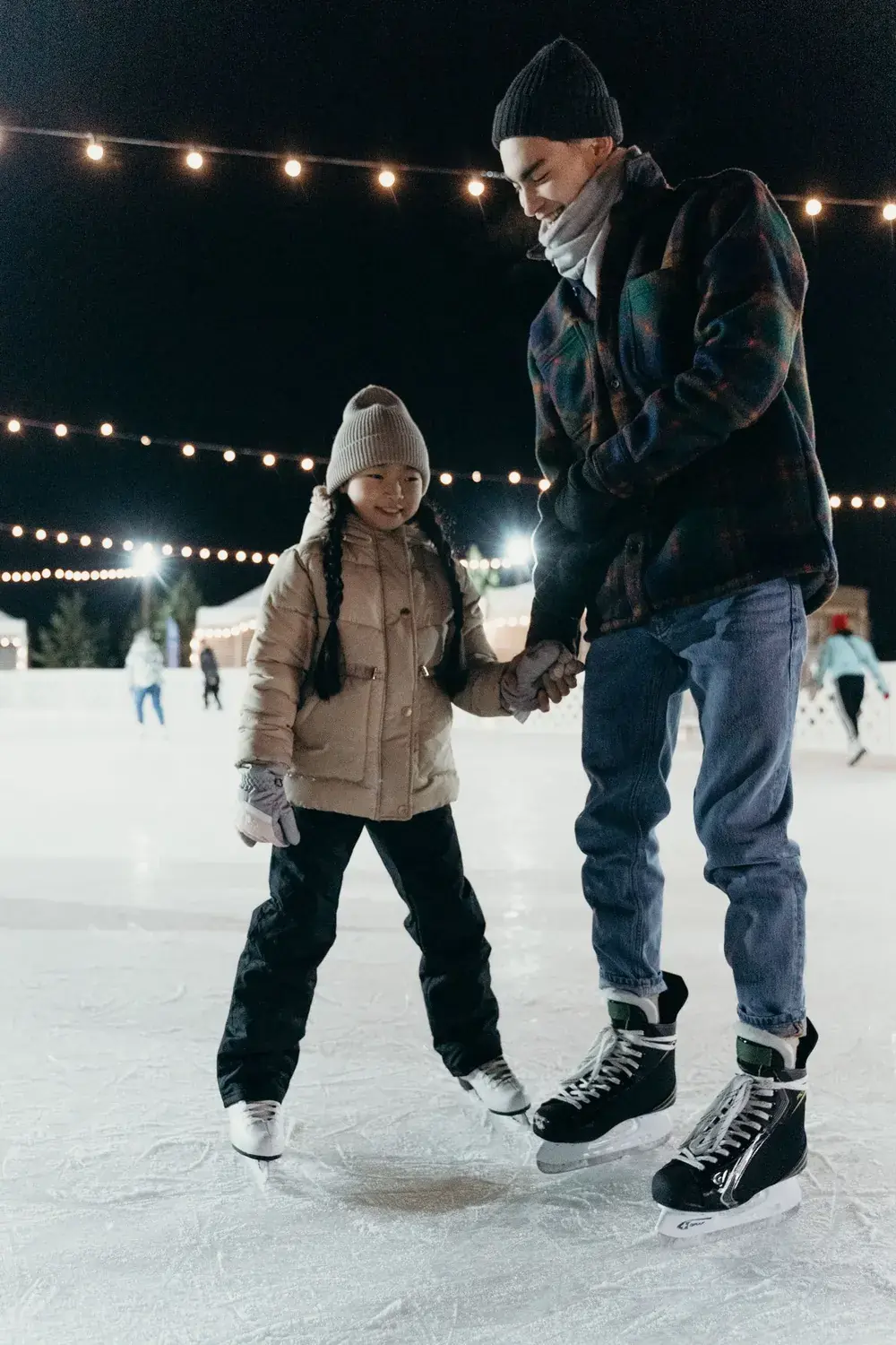A dad holding his daughter's hand as they skate under festoon lights at an evening session